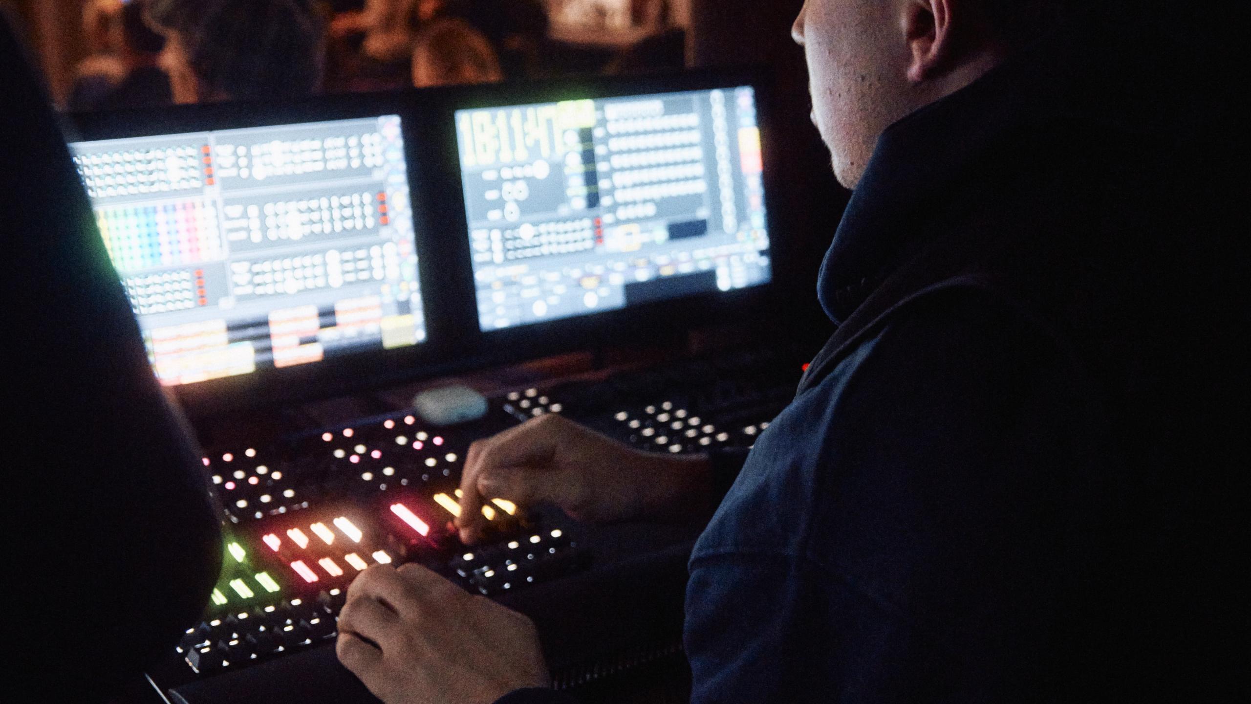 Techniker steuert Licht- und Soundanlage während einer Veranstaltung im Bergson-Kunst- und Kulturareal. Zwei Monitore mit Mischpult leuchten im Dunkeln, im Hintergrund ist das Publikum in warmer Atmosphäre zu sehen.