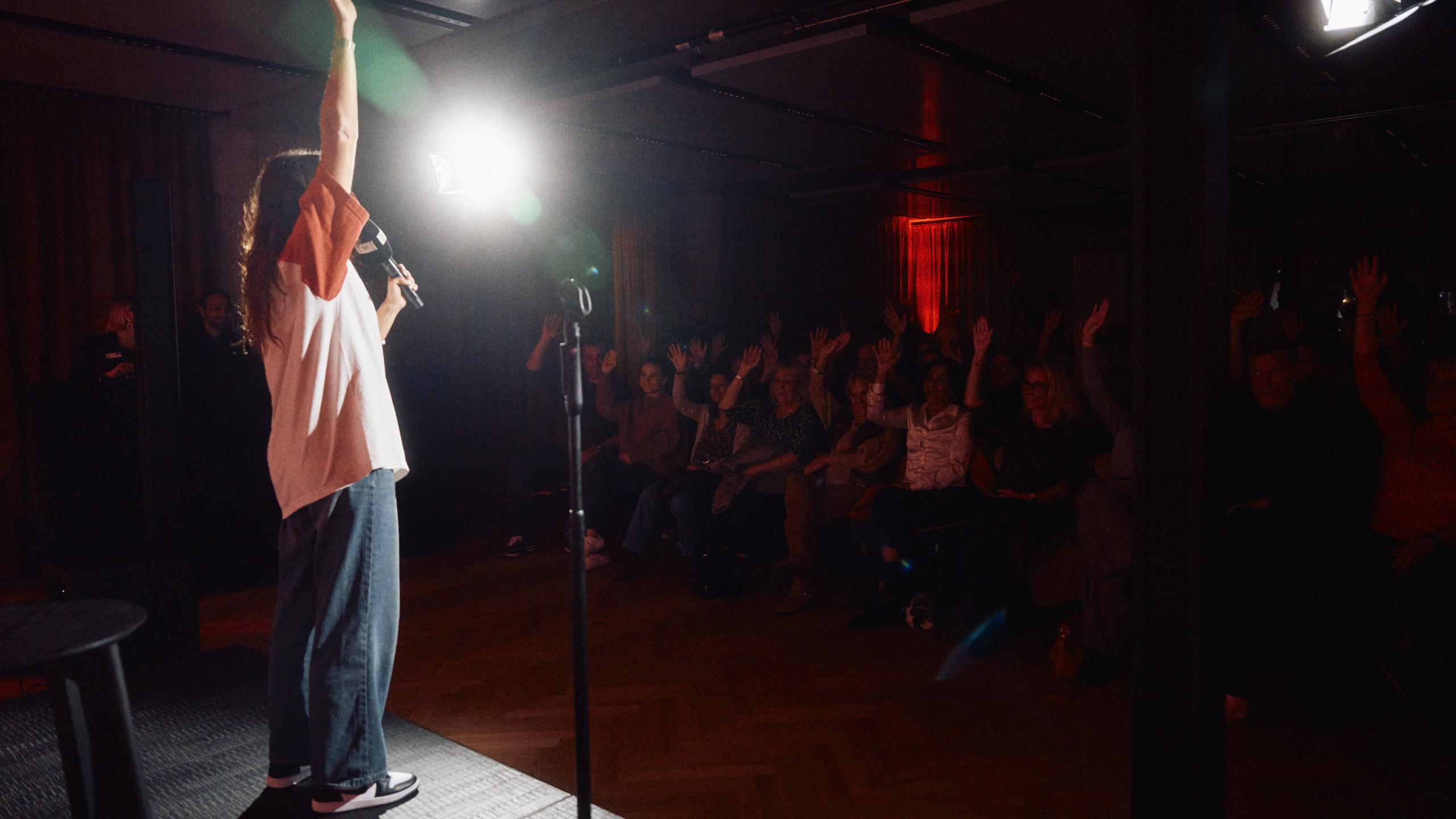 Künstlerin auf der Bühne im Bergson-Kulturareal spricht ins Mikrofon, während das Publikum mit erhobenen Händen reagiert. Warme Lichtstimmung, mitreißende Live-Atmosphäre bei einer Performance oder Lesung.