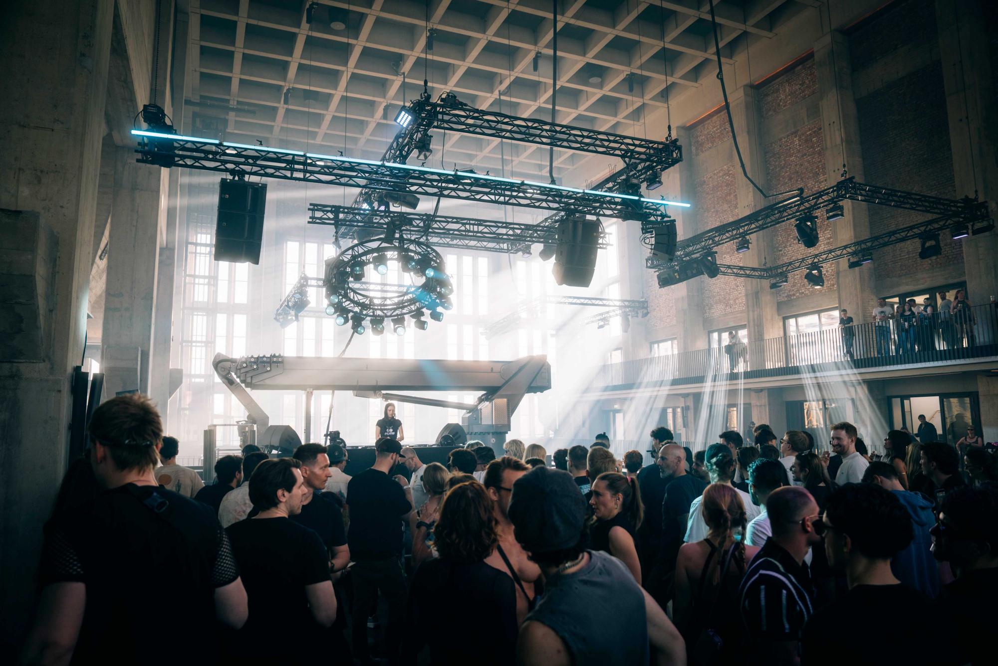 DJ-Set vor tanzender Menge in einer industriellen Eventhalle mit hohen Betondecken, Lichttraverse und einfallendem Tageslicht – Clubnacht im Bergson Kunstkraftwerk München – Foto von Simon Haseneder